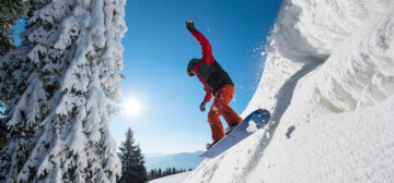 Motion picture of a snowboarder flying in the air while riding the snowy slope in winter. Blue sky, sun, forest and mountains on the background copyspace sport extreme people lifestyle concept